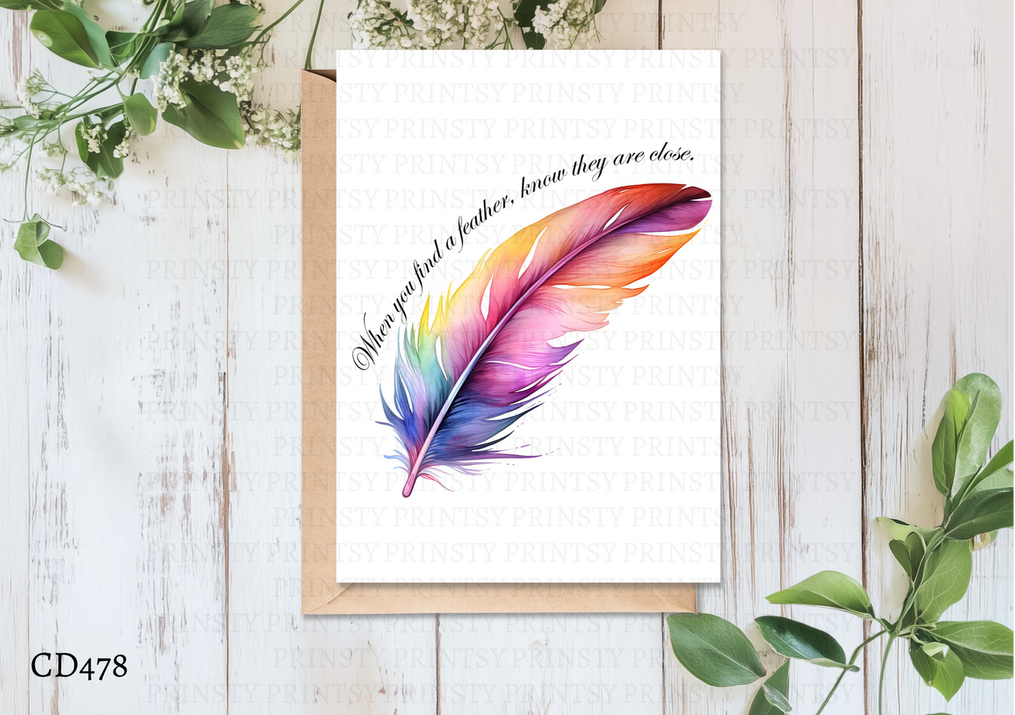 Colorful feather illustration on a card with a quote, placed on a wooden surface with greenery.