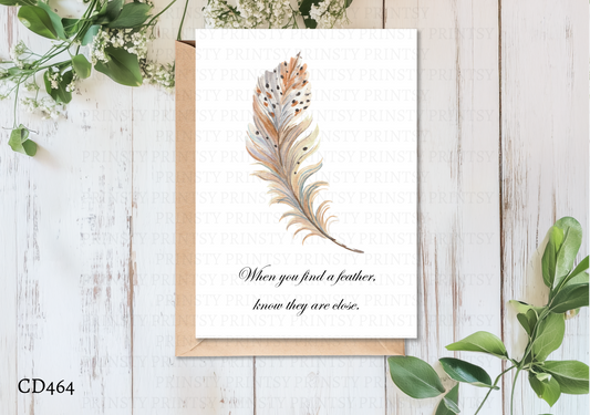 Greeting card with feather design and text on a wooden surface with greenery
