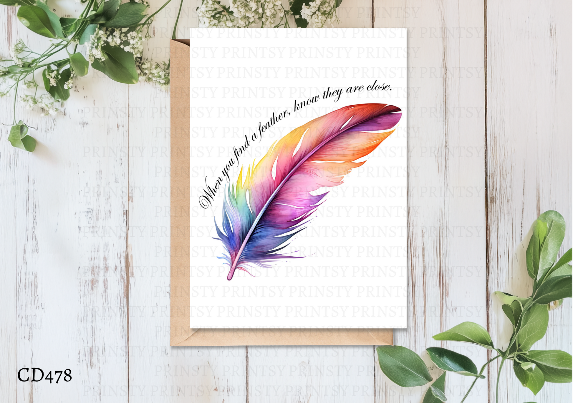 Colorful feather illustration on a card with a quote, placed on a wooden surface with greenery.